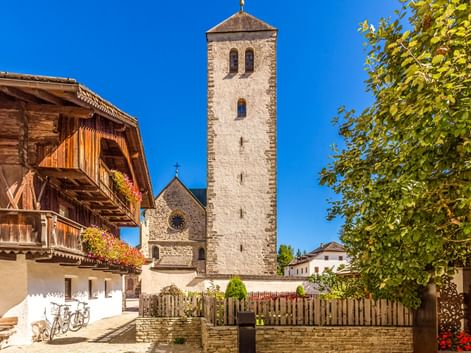 Stone bell tower of Innichen Collegiate Church rising against blue sky, with traditional Alpine wooden house and green tree framing the scene.