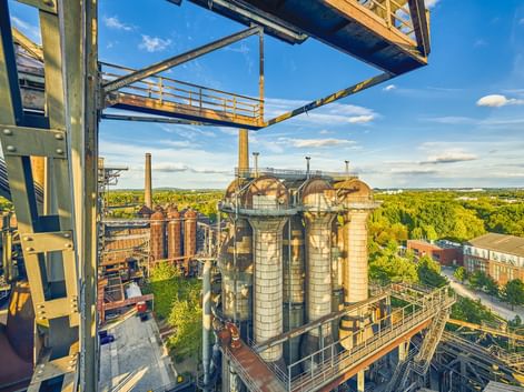 Erhöhte Ansicht historischer Hochöfen und Industriestrukturen im Landschaftspark Duisburg-Nord, umgeben von grüner Vegetation unter blauem Himmel.
