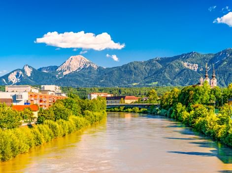 View of Villach with the Drau River flowing through green banks, colorful buildings, a church, and dramatic mountain peaks under blue sky.