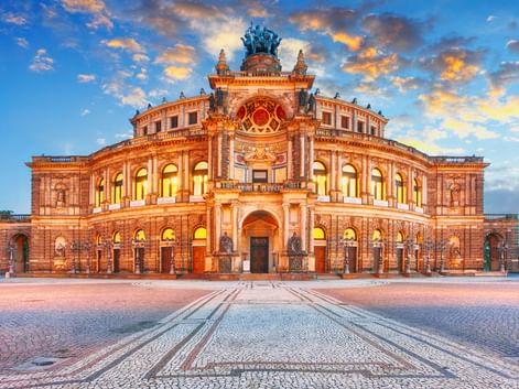 Beleuchtete Semperoper in Dresden mit prachtvoller Barockarchitektur unter dramatischem Sonnenuntergangshimmel mit orangen und blauen Wolken.