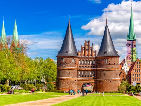 Das Holstentor, ein mittelalterliches Stadttor aus rotem Backstein mit zwei runden Türmen in Lübeck, umgeben von grünen Rasenflächen und Kirchtürmen.