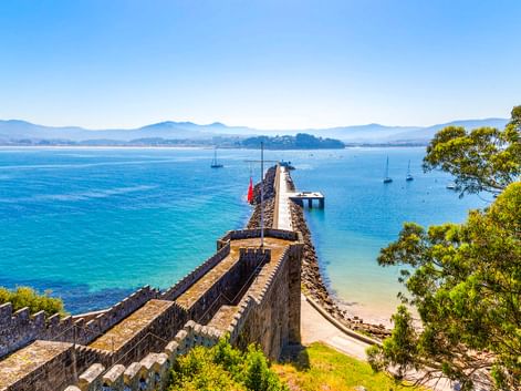 Steinmauern der Festung Monterreal erstrecken sich in die Bucht von Baiona mit Segelbooten im türkisfarbenen Wasser und Bergen in der Ferne.