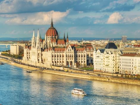 Panoramablick auf das ungarische Parlamentsgebäude am Donauufer in Budapest mit Booten auf dem Wasser und einer Brücke im Hintergrund.