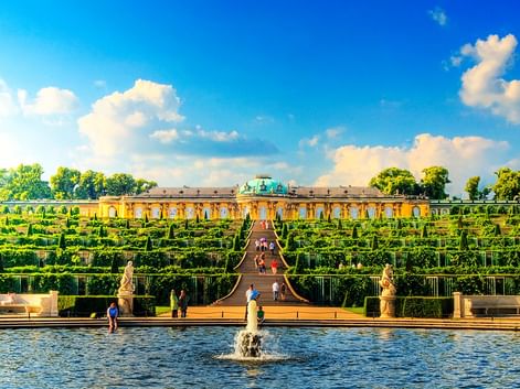 Schloss Sanssouci in Potsdam mit gelber Fassade und terrassierten Weingärten. Brunnen im Vordergrund, Besucher auf Treppe, blauer Himmel mit Wolken.