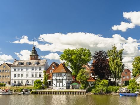 Uferblick auf Glückstadt mit historischen Gebäuden, darunter ein weißer Kirchturm, Fachwerkhaus und Boote am Hafen unter blauem Himmel.