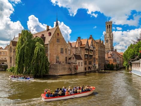 Zwei Touristenboote auf einem Kanal in Brügge mit mittelalterlichen Gebäuden und dem Belfried-Turm unter blauem Himmel mit weißen Wolken.