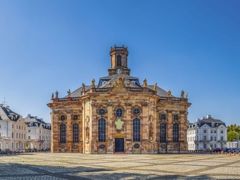 Ludwigskirche, eine barocke Kirche mit gelber Sandsteinfassade und Turm, auf einem Kopfsteinpflasterplatz in Saarbrücken unter blauem Himmel.