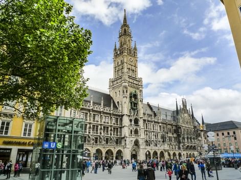 Marienplatz in München mit dem gotischen Rathausturm vor blauem Himmel. Fußgänger versammeln sich auf dem Platz mit grünen Bäumen links.