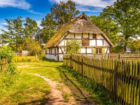 Fachwerkhaus mit Reetdach im Fischerdorf Kluki, umgeben von Holzzaun und grünen Bäumen unter blauem Himmel.
