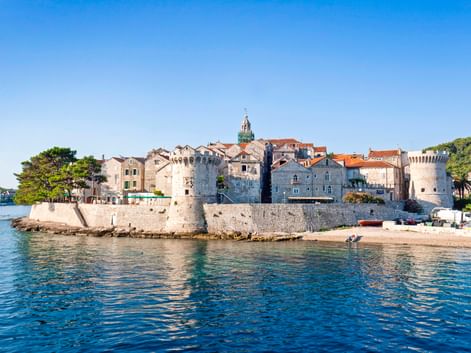 Blick auf die Altstadt von Korcula mit steinernen Befestigungsmauern, historischen Gebäuden mit Terrakottadächern und Kirchturm unter blauem Himmel.
