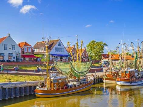 Traditionelle Fischerboote mit Netzen liegen im Hafen von Greetsiel. Bunte Häuser mit roten Dächern säumen das Ufer unter blauem Himmel.