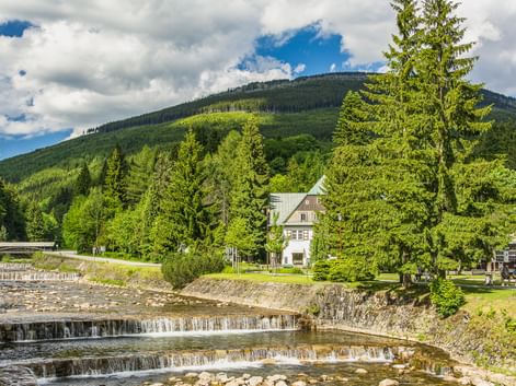 Kaskadenartige Stromschnellen der Labe in Spindlermühle mit Steinterrassen, umgeben von grünen Bäumen und einem weißen Haus, Berge im Hintergrund.