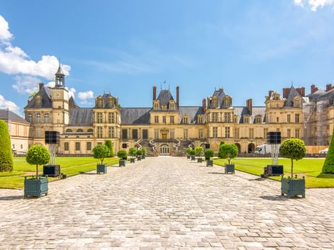 Fassade des Schlosses Fontainebleau mit Kopfsteinpflaster-Innenhof, gepflegten Rasenflächen und Formschnittbäumen unter blauem Himmel.
