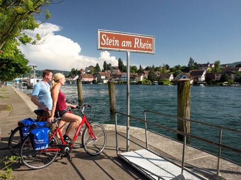 Zwei Radfahrer mit rotem Fahrrad am Ortsschild Stein am Rhein am Rheinufer. Historische Stadtgebäude sichtbar über dem türkisfarbenen Wasser.