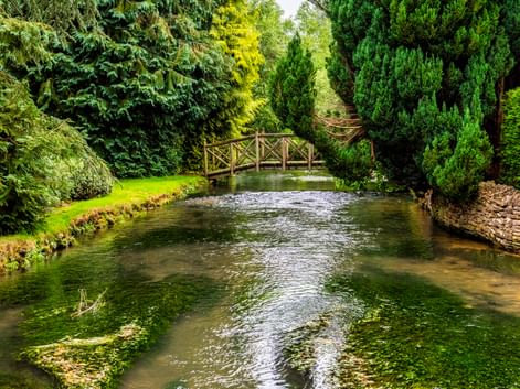 Klarer Bach Fluss Windrush fließt durch Bourton-on-the-Water mit Holzbrücke, üppigen grünen Bäumen und grasbewachsenen Ufern an einem sonnigen Tag.