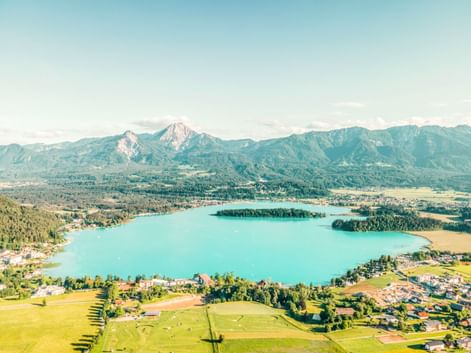 Luftaufnahme vom türkisfarbenen Faaker See in Österreich mit bewaldeten Inseln, grünen Wiesen und Bergkette im Hintergrund unter klarem Himmel.