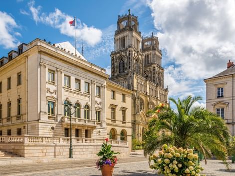 Gotische Zwillingstürme der Kathedrale Sainte-Croix in Orléans hinter einem cremefarbenen Gebäude mit Palmen und Blumen auf dem Platz.