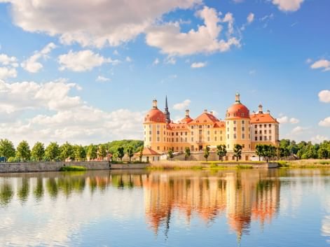 Moritzburg Castle with yellow facade and red domes reflected in surrounding lake under blue sky with white clouds.