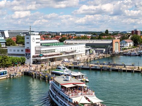 Fahrgastschiff im Friedrichshafener Hafen am Bodensee. Weiße Hafengebäude und Holzstege unter blauem Himmel mit weißen Wolken.