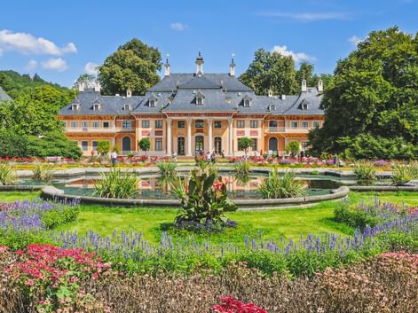 Schloss Pillnitz mit oranger Fassade und grauem Dach hinter Zierbrunnen und bunten Blumenbeeten im barocken Garten unter blauem Himmel.
