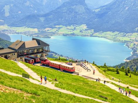Rote Zahnradbahnen an der Schafberg-Bergstation mit Blick auf türkisfarbenen See und grüne Täler mit Alpengipfeln im Hintergrund.