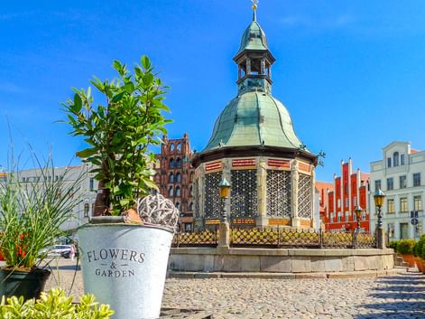 Verzierter Pavillon mit Kupferkuppel auf Kopfsteinpflasterplatz in Wismar. Bunte historische Gebäude im Hintergrund unter blauem Himmel.