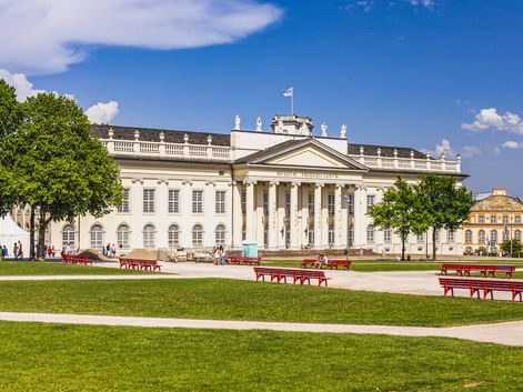 The neoclassical Fridericianum building in Kassel with columned facade, viewed across a green lawn with red benches under blue sky.