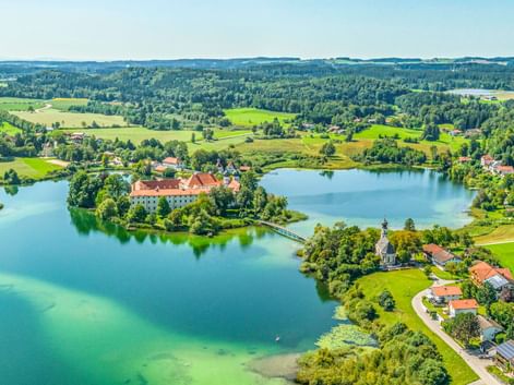 Luftaufnahme des Klosters Seeon auf einer Halbinsel in einem türkisfarbenen See, umgeben von grünen Feldern, Wäldern und einem Dorf in Bayern.