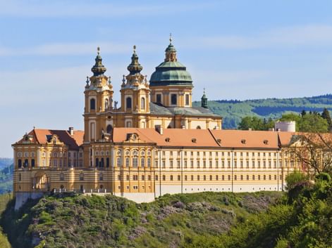 Melk Abbey perched on a rocky hill in Austria. The baroque monastery features yellow-cream facades, twin towers, and a green dome against blue sky.