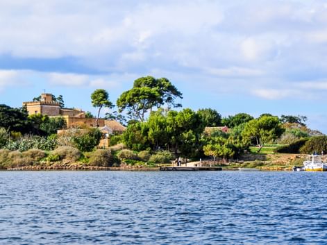 Blick auf Mozia auf der Insel San Pantaleo mit historischen Gebäuden, üppigen grünen Bäumen und blauem Wasser im Vordergrund unter bewölktem Himmel.
