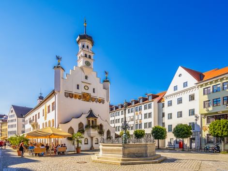 Historisches weißes Kemptener Rathaus mit Glockenturm auf Kopfsteinpflasterplatz. Brunnen im Vordergrund, bunte Gebäude und Café unter blauem Himmel.