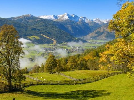 Alpental bei St. Michael im Lungau mit grünen Wiesen, Holzzäunen, Herbstbäumen und schneebedeckten Bergen unter Morgennebel.