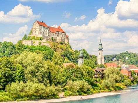 Burg Burghausen mit roten Dächern auf einem Hügel über einer Stadt mit Kirchtürmen und grünem Flussufer. Blauer Himmel mit weißen Wolken.