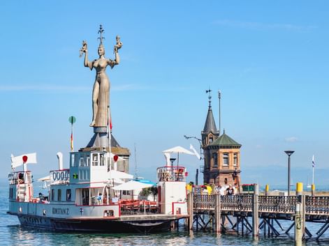 Imperia-Statue am Konstanzer Hafen mit Fahrgastschiff und historischem Turm. Die hohe rotierende Statue steht auf einem Sockel am Wasser.