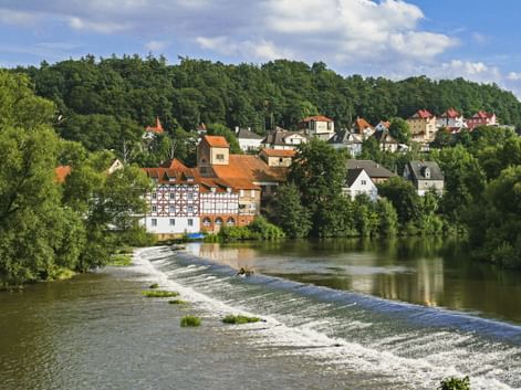 View of Melsungen on the Fulda River with a weir in the foreground. Half-timbered houses and red roofs nestle among green trees under a blue sky.