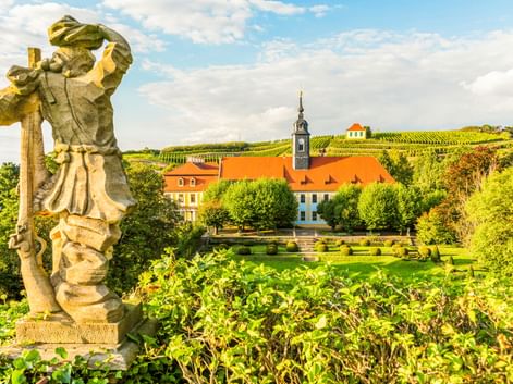 Stone statue in foreground overlooking gardens and historic buildings with orange roofs in Diesbar-Seußlitz, with vineyard-covered hills behind.