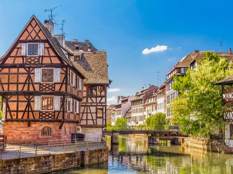 Traditionelle Fachwerkhäuser entlang eines Kanals in La Petite France, Straßburg, mit Brücke und grünen Bäumen unter blauem Himmel.