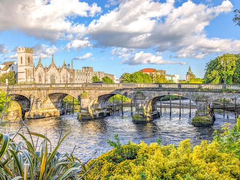 Historische steinerne Salmon Weir Bridge mit fünf Bögen über dem River Corrib in Galway, Irland. Gotische Kathedrale und üppiges Grün rahmen die Szene.