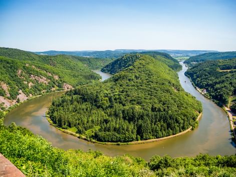 Luftaufnahme der Saarschleife, einer hufeisenförmigen Flussbiegung, umgeben von dicht bewaldeten Hügeln unter blauem Himmel.