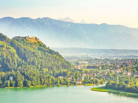 Burgruine Landskron auf einem bewaldeten Hügel mit Blick auf Villach und einen türkisfarbenen See, mit Bergketten im Hintergrund bei weichem Licht.