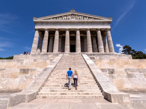 Zwei Personen steigen die Steinstufen zur Walhalla hinauf, einem neoklassizistischen Tempel mit dorischen Säulen in Bayern, unter blauem Himmel.