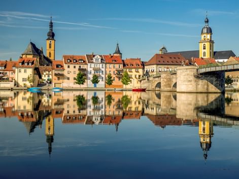 Kitzinger Altstadt mit bunten Gebäuden und Kirchtürmen, die sich im ruhigen Main spiegeln. Rechts überspannt eine Steinbrücke das Wasser.