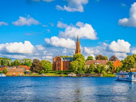 Blick über den Malchower See mit Fahrgastschiff auf blauem Wasser. Backsteinkirche mit hohem Turm ragt über Ufergebäude unter blauem Himmel.