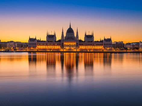 Beleuchtetes ungarisches Parlamentsgebäude am Donauufer bei Sonnenuntergang mit goldenen Lichtern, die sich im ruhigen Wasser spiegeln.