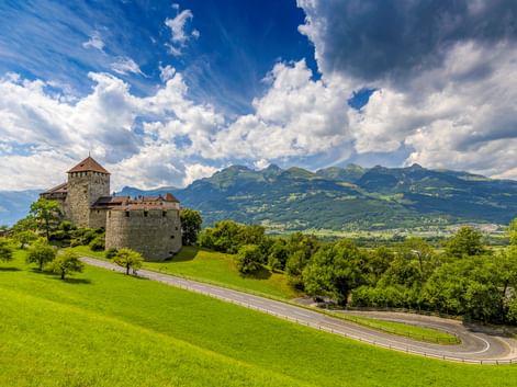 Schloss Vaduz auf einem grünen Hügel mit kurvenreicher Straße, umgeben von Bäumen. Alpenberge und dramatische Wolken im Hintergrund.