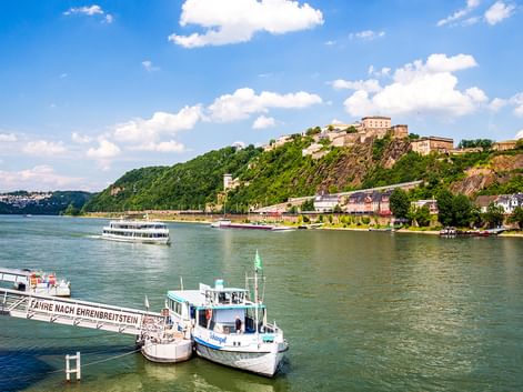 Festung Ehrenbreitstein auf einem Hügel über dem Rhein in Koblenz. Fähren und Passagierschiffe auf dem Wasser, blauer Himmel mit Wolken.