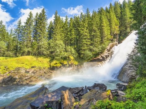 Krimmler Wasserfälle stürzen über Felsen in türkisfarbenes Becken, umgeben von grünem Wald und Wiesen unter blauem Himmel mit aufsteigendem Nebel.