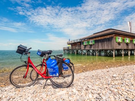 Rotes Tourenrad mit blauen Taschen am Kieselstrand in Bregenz. Hölzerne Badehütte auf Stelzen mit grünen Fensterläden ragt in den Bodensee.