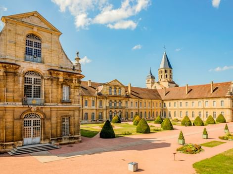 Abtei von Cluny mit barocker Kirchenfassade, langem gelbem Klostergebäude und formalen Gärten mit konischen Hecken unter blauem Himmel.