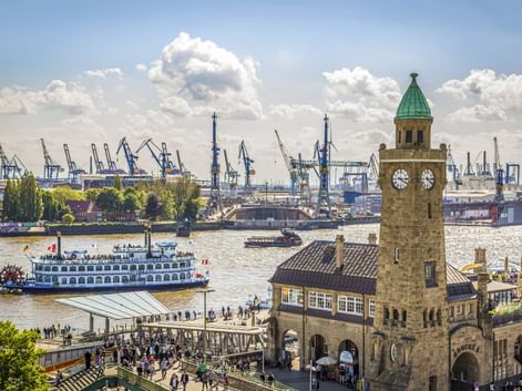 St. Pauli Landungsbrücken in Hamburg mit Uhrturm, Raddampfer auf der Elbe und Hafenkränen im Hintergrund unter bewölktem Himmel.